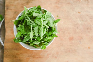 Beyaz bir kâseye odaklanan tepeden çekimler. Açık ahşap bir yüzeye yerleştirilmiş taze yeşil yapraklarla dolu..