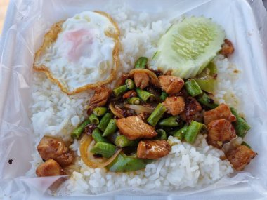 Boxed lunch ready to eat. Cooked rice with chicken chilli, fried egg and cucumber in a foam box lined with clear plastic sheet.