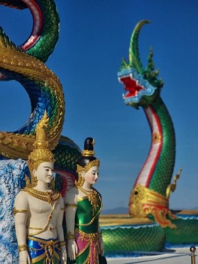 İki süslü tanrı görkemli çok başlı Naga yılanlarının önünde duruyor. Efsanevi yaratıklar açık mavi gökyüzüne karşı canlı yeşil ve altın pullar sergiliyor. Bu karmaşık heykel ruhani sanatları sergiliyor..