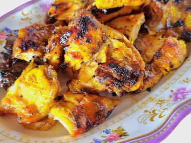 Kızarmış tavuk parçalarının yakın çekimi, çıtır çıtır derisi ve görünen ızgara izleri çiçeklerle süslenmiş beyaz bir tabakta servis ediliyor..