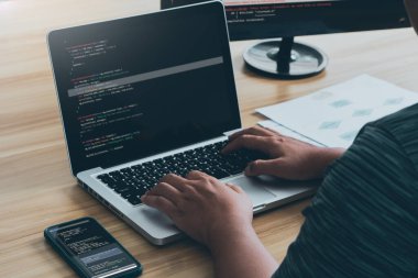 Young programmer writing program code sitting at the workplace with laptop and mobile phone in the office.