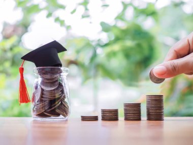 Mezuniyet şapkası takıp bozuk para ve cam şişeye para koymak için elini kapat. Eğitim için para biriktirme kavramı, öğrenci kredisi, burs, gelecek eğitim ücretleri