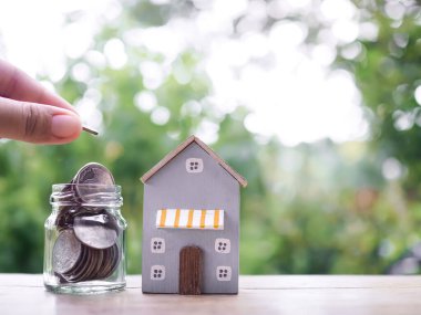 Kapalı el, cam şişe ve minyatür eve bozuk para koyuyor. Ev için para biriktirme konsepti, emlak yatırımları, ev kredisi, gayrimenkul..