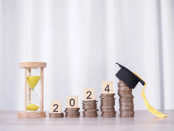 Study goals, Hourglass and Wooden block with number 2024 on stack of coins with graduation hat. The concept of saving money for education, student loan, scholarship, tuition fees in New Year 2024