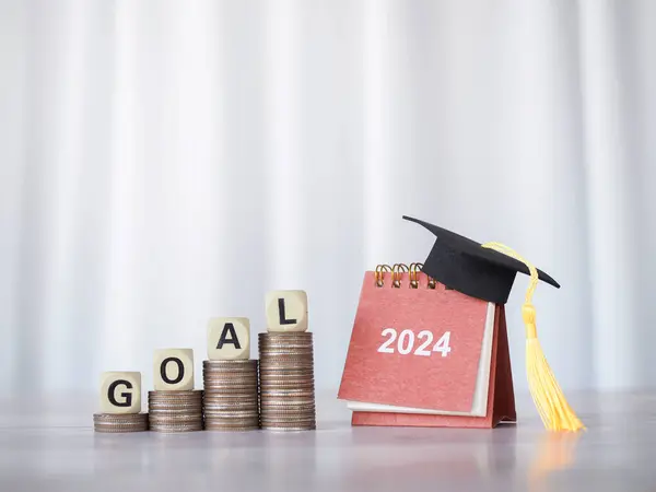 Study goal, 2024 desk calendar with graduation hat and Wooden block with word GOAL on stack of coins. The concept of saving money for education, student loan, scholarship, tuition fees in Year 2024