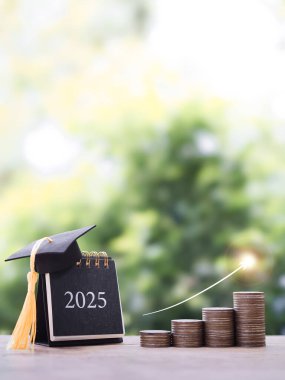 Mezuniyet şapkalı 2025 masa takvimi, yükselen bir yığın bozuk para ve ok. 2025 yılında eğitim, öğrenci kredisi, burs, öğrenim ücreti için para biriktirme kavramı