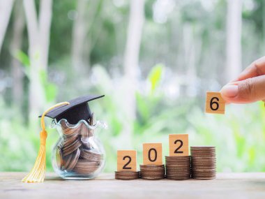 Mezuniyet şapkalı para yığınının üzerinde 2026 numaralı tahta bloklar var. 2026 yılında eğitim, öğrenci kredisi, burs, öğrenim ücreti için para biriktirme kavramı