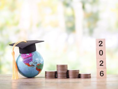 Mezuniyet şapkalı dünya küresi, 2026 numaralı tahta blok ve bir yığın bozuk para. 2026 yılında eğitim, öğrenci kredisi, burs, öğrenim ücreti için para biriktirme kavramı.