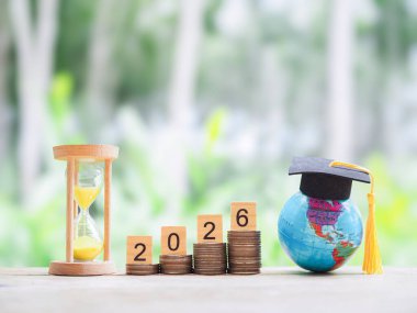 Hedeflere çalış. Kum saati, Dünya küresi, mezuniyet şapkası ve bozuk para yığını. 2026 yılında eğitim, öğrenci kredisi, burs, öğrenim ücreti için para biriktirme kavramı