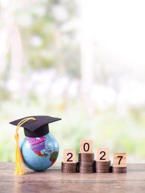 Mezuniyet şapkalı dünya küresi, 2027 numaralı tahtadan blok madeni para yığını. 2027 yılında eğitim, öğrenci kredisi, burs, öğrenim ücreti için tasarruf kavramı.
