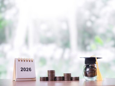 Hedeflere çalış. 2026 masa takvimi, mezuniyet şapkalı cam şişe ve bozuk para yığını. 2026 yılında eğitim, öğrenci kredisi, burs, öğrenim ücreti için para biriktirme kavramı