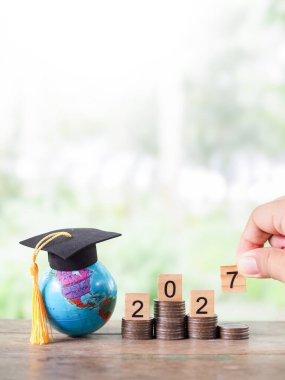 Mezuniyet şapkalı dünya küresi, 2027 numaralı tahtadan blok madeni para yığını. 2027 yılında eğitim, öğrenci kredisi, burs, öğrenim ücreti için tasarruf kavramı.