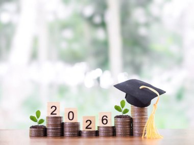 Mezuniyet şapkalı para yığınının üzerinde 2026 numaralı tahta bloklar var. 2026 yılında eğitim, öğrenci kredisi, burs, öğrenim ücreti için para biriktirme kavramı