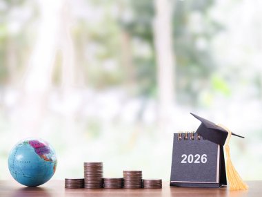 Dünya küresi, 2026 mezuniyet şapkası ve bozuk paralarla dolu masa takvimi. 2026 yılında eğitim, öğrenci kredisi, burs, öğrenim ücreti için para biriktirme kavramı