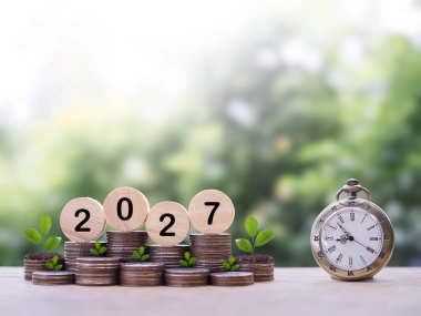Kaliteli saat, 2027 numaralı tahta bloklar ve bozuk para yığınında büyüyen bitkiler. 2027 yılında başarılı olmak için zaman kazanma kavramı