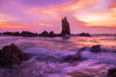 Gün batımındaki büyük taşların görüntüsü, su sıçramalarıyla kayaların üzerinde kırılan deniz dalgaları.