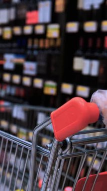 Bir müşteri, şarap şişeleriyle dolu bir arabayı iterek dolu bir şarap dükkanında geziniyor. Sahnede çeşitli kırmızılar, beyazlar ve köpüklü şaraplar, bolluk ve seçim duyguları sergileniyor.