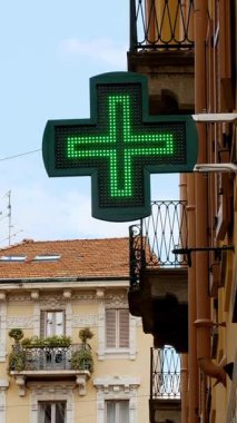 A vibrant green pharmacy cross sign stands out against an urban backdrop, nestled between traditional european architecture with Balconies and weathered facades under a clear sky
