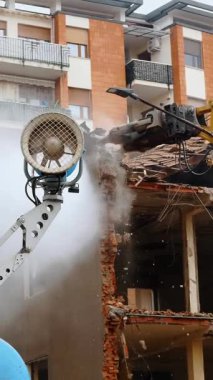 Hidrolik kollu bir kazıcı, yaşlanmakta olan tuğla bir binayı yıkıp havaya toz bulutu göndererek kentsel gelişimi ve yenilenmeyi sembolize ediyor.