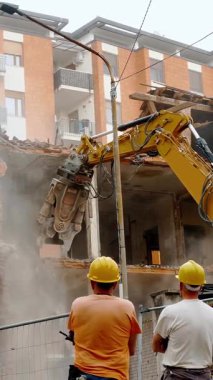 Professional construction workers in hard hats observe an excavator demolishing a building, ensuring safety and efficiency amidst rising dust in an urban environment