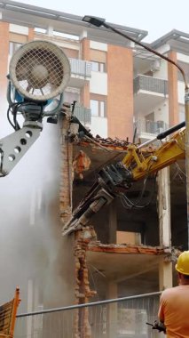 Workers in safety helmets observe as a heavy excavator operates on a demolition site, tearing down parts of an old building amidst urban surroundings