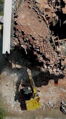 Yıllanmış tuğla bina, yıkım ve inşaat endüstrisinin yıkıcı faaliyetleri arasında sarı bir kazıcıyı gösteren hava görüntüsü