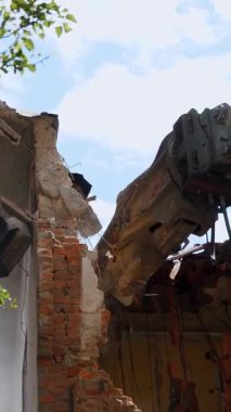 Zarar görmüş tuğla bir binanın kalıntılarını parçalayan sarı kazıcının detaylı bir görüntüsü, açık mavi gökyüzüne karşı yeşil yapraklar arasında.