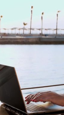 Koruyucu maskeli genç bir kadın, güneş şapkası ve yaz kıyafetleri, dizüstü bilgisayarda çalışıyor, deniz kenarındaki boş bir barajda oturuyor. Uzaktan çalış. Covid-19 'un peşinden gidiyorum. yaz tatili.