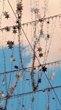 Yakından bakınca, ampullerden ve gümüş yıldızlardan oluşan çelenkler rüzgarda sallanıyor, güneş ışınlarında parıldıyor. Mavi gökyüzüne karşı hafif sokak dekorasyonu.