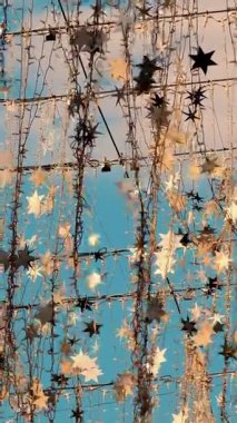 Yakından bakınca, ampullerden ve gümüş yıldızlardan oluşan çelenkler rüzgarda sallanıyor, güneş ışınlarında parıldıyor. Mavi gökyüzüne karşı hafif sokak dekorasyonu.