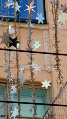 Yakından bakınca, ampullerden ve gümüş yıldızlardan oluşan çelenkler rüzgarda sallanıyor, güneş ışınlarında parıldıyor. Hafif sokak dekorasyonu