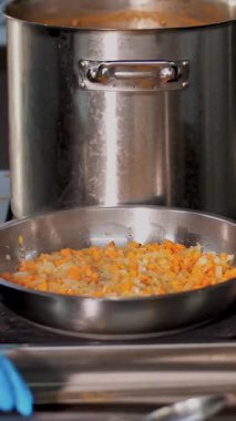 Kantinde yemek pişirmek. Yakın plan. Ocakta büyük tencereler var. Soğan ve havuç tavada kızartılır. Aşçı, koruyucu eldivenler içinde, kızartmayı karıştırıyor, ve sonra da çorba. Sağlıklı gıda ve güvenlik kavramı. yeniden açılıyor