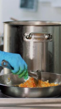 Kantinde yemek pişirmek. Yakın plan. Ocakta büyük tencereler var. Soğan ve havuç tavada kızartılır. Aşçı, koruyucu eldivenlerle, kızartmayı karıştırıyor. Sağlıklı gıda ve güvenlik kavramı. yeniden açılıyor