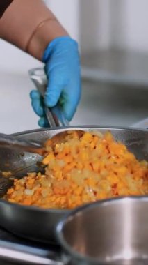 Yemek pişirmek. Yakın plan. Aşçı koruyucu eldivenlerle, kızartma tavasında kızarmış soğan ve havuçları karıştırıyor. Sağlıklı gıda ve güvenlik kavramı. Matarayı yeniden aç
