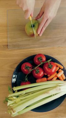 İnternette yemek yapmak. Sağlıklı yemek. Yakın plan. Üst Manzara. Kadın, ahşap masada cam tahtada elma kesiyor. Tabakta çeşitli sebze ve meyveler var. Mutfak vlogları, sosyal ağlar.