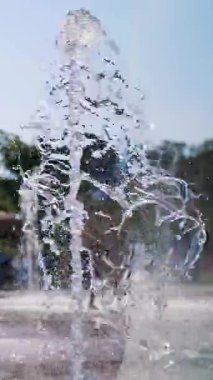 Çeşme. Yakın plan. Fıskiye fıskiyeleri. Arka planda bulanık çocuklar, su akıntıları arasında eğleniyorlar, şehir parkında yüksek çeşme jetleri, sıcak yaz günü.