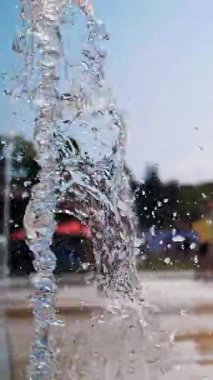 Çeşme. Yakın plan. Fıskiye fıskiyeleri. Arka planda bulanık çocuklar, su akıntıları arasında eğleniyorlar, şehir parkında yüksek çeşme jetleri, sıcak yaz günü.