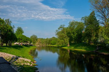 Beautiful colorful summer or spring natural landscape with a lake in a park surrounded by green foliage of trees in sunlight and a stone path in the foreground.