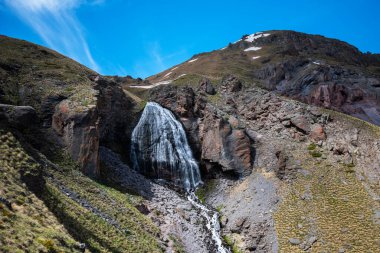 A mountain stream flows down from the rocks with melted snow in the high mountains. Green vegetation in the mountains in summer.