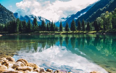Yaz, gün ışığında dağları yeşil gölün sakin sularına yansıtır. Dağ manzarası, göl ve dağ sırası, geniş bir manzara..
