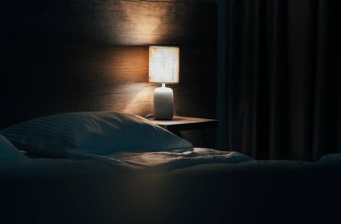 The cozy bed in the hotel at night with yellow night lamp. The light from the lamp reflects on the wooden wall. Toned image with copy space for you text. clipart