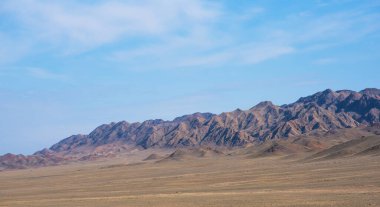 Geniş, düz ovaları olan geniş bir çöl, seyrek, kuru bitki örtüsü ve yuvarlanan tepelerle kaplı, güneşin ışığıyla engebeli bir dağ sırasına uzanan, gölgeler oyunu yaratan,.