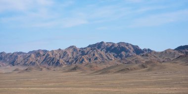 Geniş, düz ovaları olan geniş bir çöl, seyrek, kuru bitki örtüsü ve yuvarlanan tepelerle kaplı, güneşin ışığıyla engebeli bir dağ sırasına uzanan, gölgeler oyunu yaratan,.