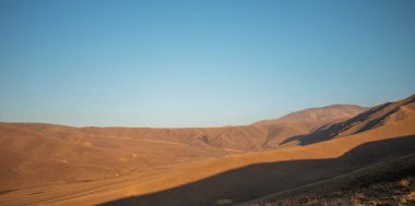 Gün batımı ışığı, yumuşak bir gökyüzünün altında gölgeli dağ sırtları ile, otlarla kaplı dağların pürüzsüz, dalgalı hatları üzerinde sıcak bir parıltı bırakır.