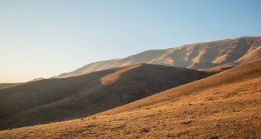 Gün batımı ışığı, yumuşak bir gökyüzünün altında gölgeli dağ sırtları ile, otlarla kaplı dağların pürüzsüz, dalgalı hatları üzerinde sıcak bir parıltı bırakır.