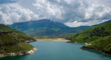 Dağlarla çevrili dağ gölünün nefes kesici manzarası. Pofuduk bulutlu ve yeşil çimen yamaçlı parlak mavi gökyüzü manzarası. Güzellik dünyası.