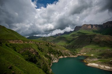 Dağlarla çevrili dağ gölünün nefes kesici manzarası. Pofuduk bulutlu ve yeşil çimen yamaçlı parlak mavi gökyüzü manzarası. Güzellik dünyası.