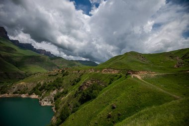 Dağlarla çevrili dağ gölünün nefes kesici manzarası. Pofuduk bulutlu ve yeşil çimen yamaçlı parlak mavi gökyüzü manzarası. Güzellik dünyası.
