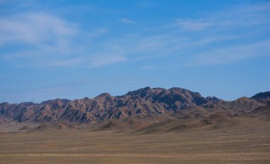 a rugged mountain range under a vast blue sky, with undulating hills in the foreground and sharp, rocky peaks in the distance creating a dramatic landscape