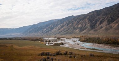 Landscape during autumn. In the foreground, grassland and meandering river bordered by trees with autumnal foliage and field with sparse vegetation. Mountains obscured by haze on background